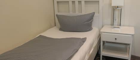 Uma cama de solteiro com cabeceira branca e travesseiro cinza, uma mesa de cabeceira com abajur e uma tomada.
