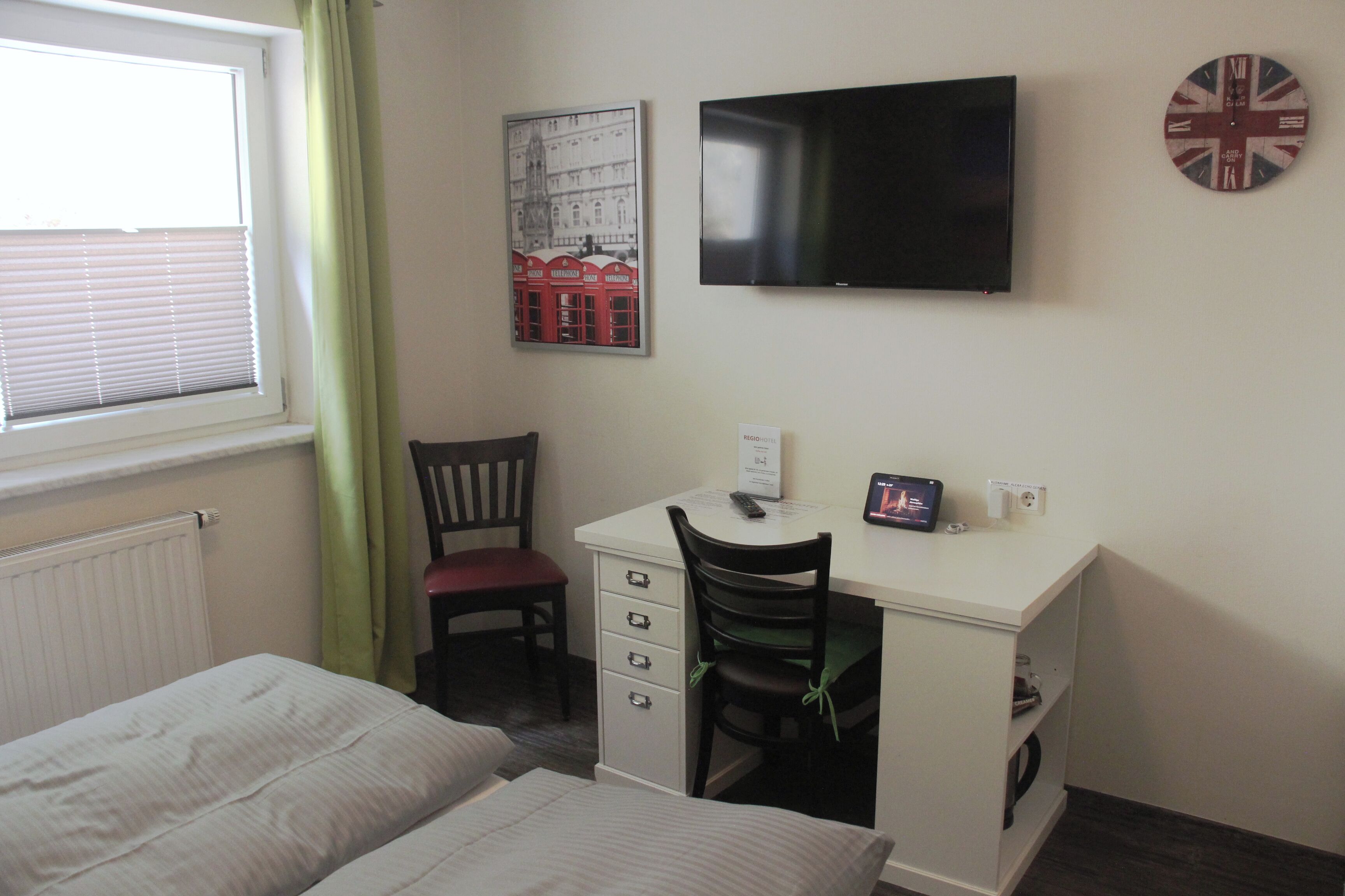 Ein Zimmer mit einem Bett, einem Schreibtisch mit Stuhl, einem Flachbildfernseher, einem gerahmten Bild und einer Wanduhr.
