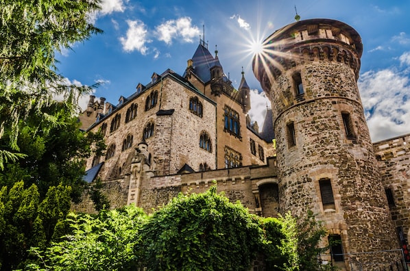 A historic stone building with multiple towers and arched windows, surrounded by lush greenery.