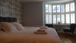 Une chambre avec un grand lit, une tête de lit ornée d’une fresque représentant une forêt, une fenêtre avec des stores, une petite table sur laquelle se trouve un bol, et une chaise.