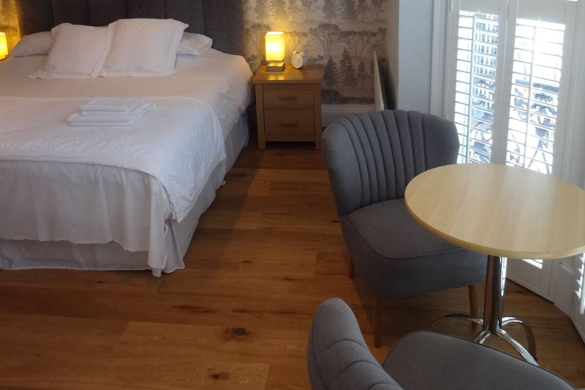 A hotel room with a bed, bedside table, lamp, chair, and a window with a view of buildings.