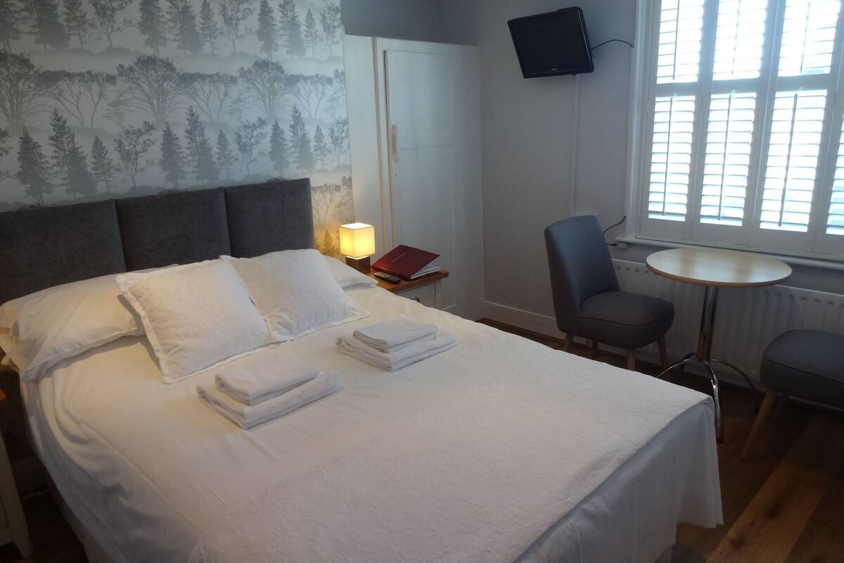 A neatly made bed with white linens and towels, a grey upholstered headboard, a small bedside table with a lamp, a chair, and a round table.
