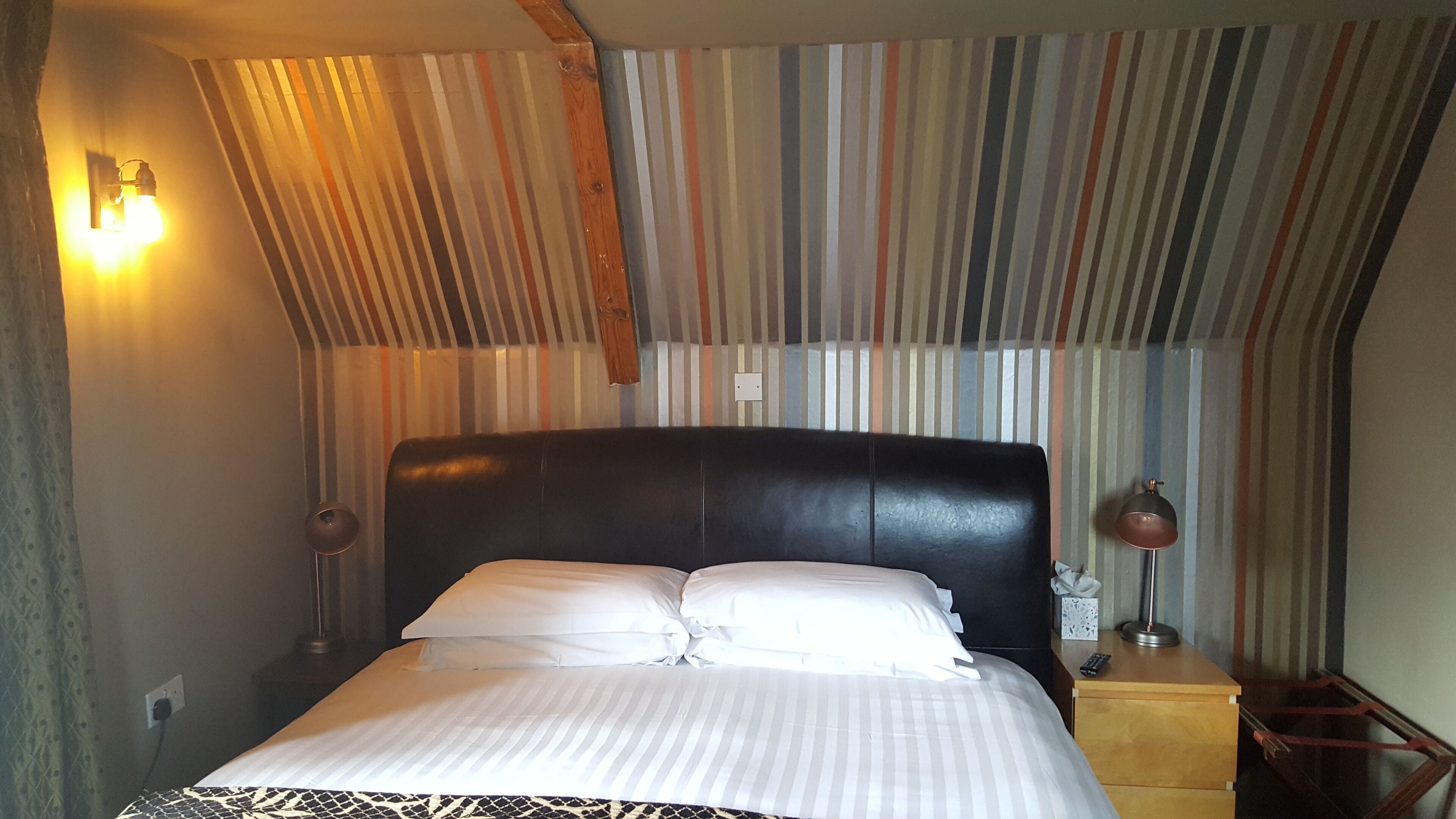A hotel room with a striped wall, a black headboard, a bed with white linens, a bedside lamp, and a wooden nightstand.
