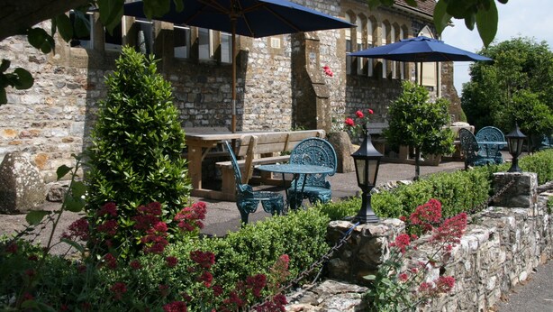 An outdoor seating area with stone walls, wooden benches, and blue umbrellas. - The Belfry at Yarcombe (Honiton)