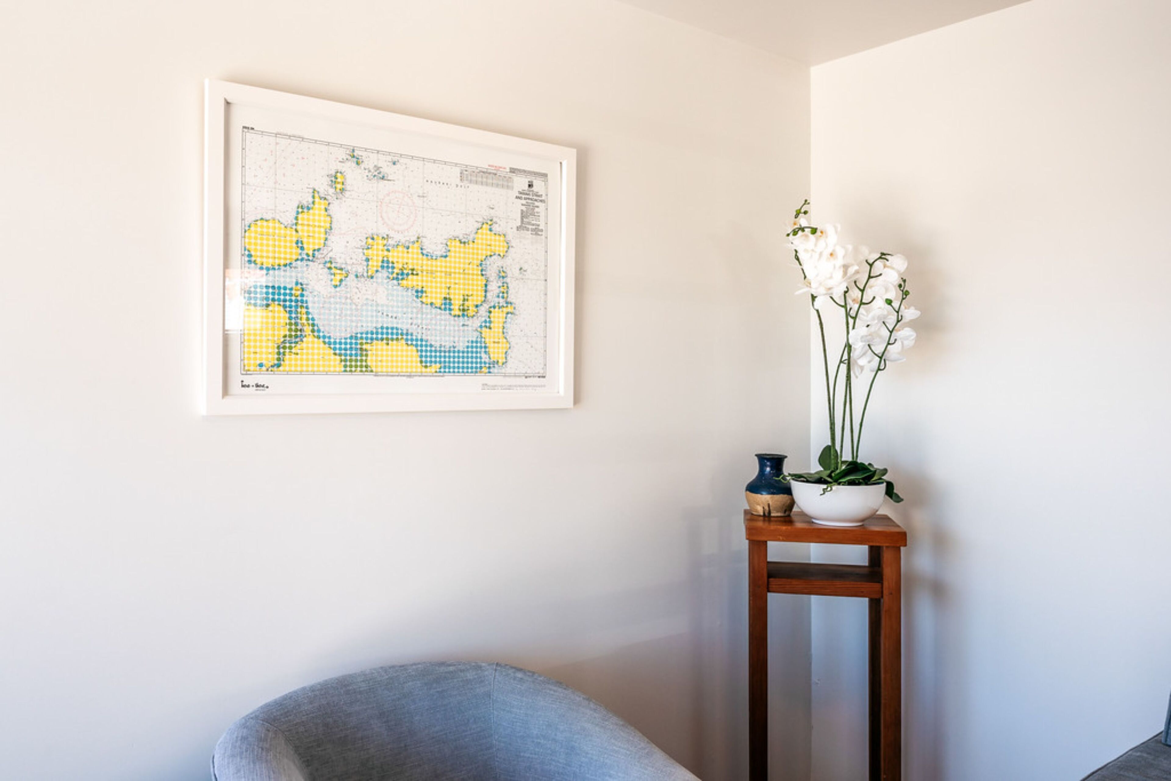 A framed map on a wall, a small wooden table with a plant, and a grey chair.