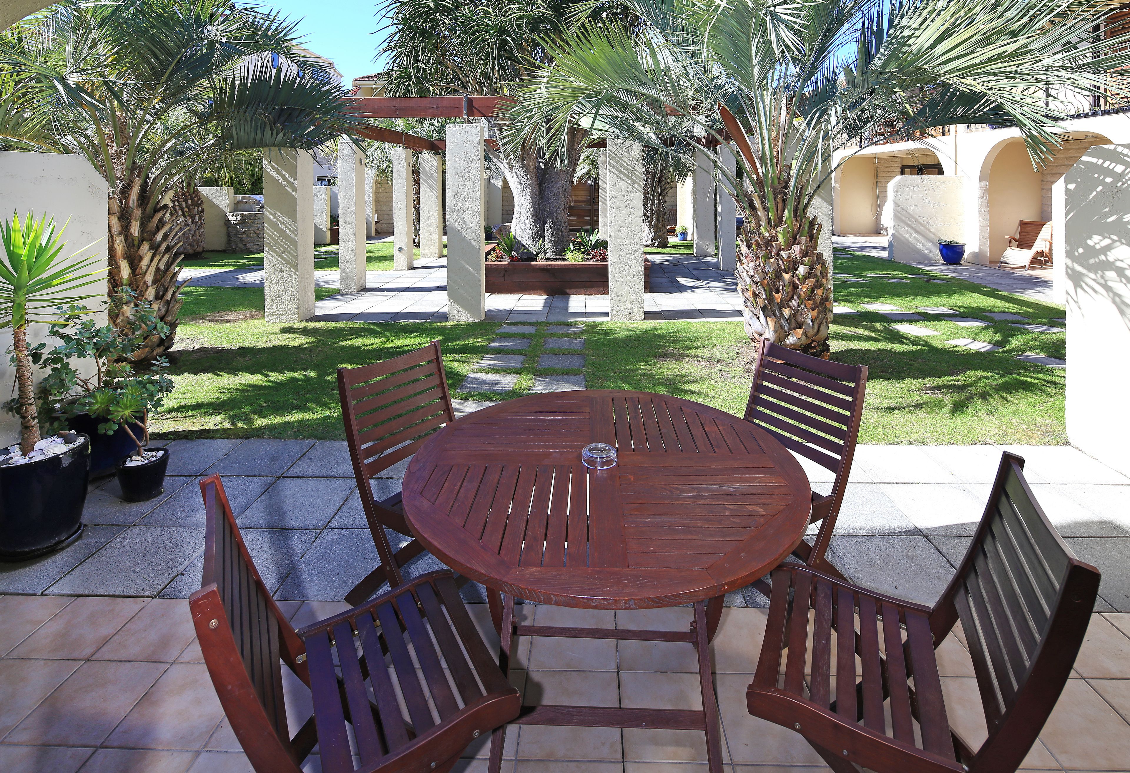 A patio with a round wooden table and four chairs, surrounded by palm trees and a stone pathway.