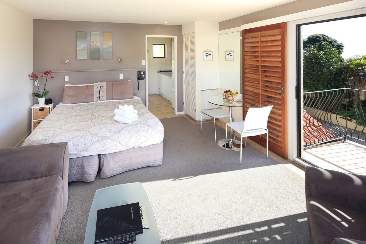 A modern bedroom with a large bed, a glass table, and a balcony with a view of the garden.