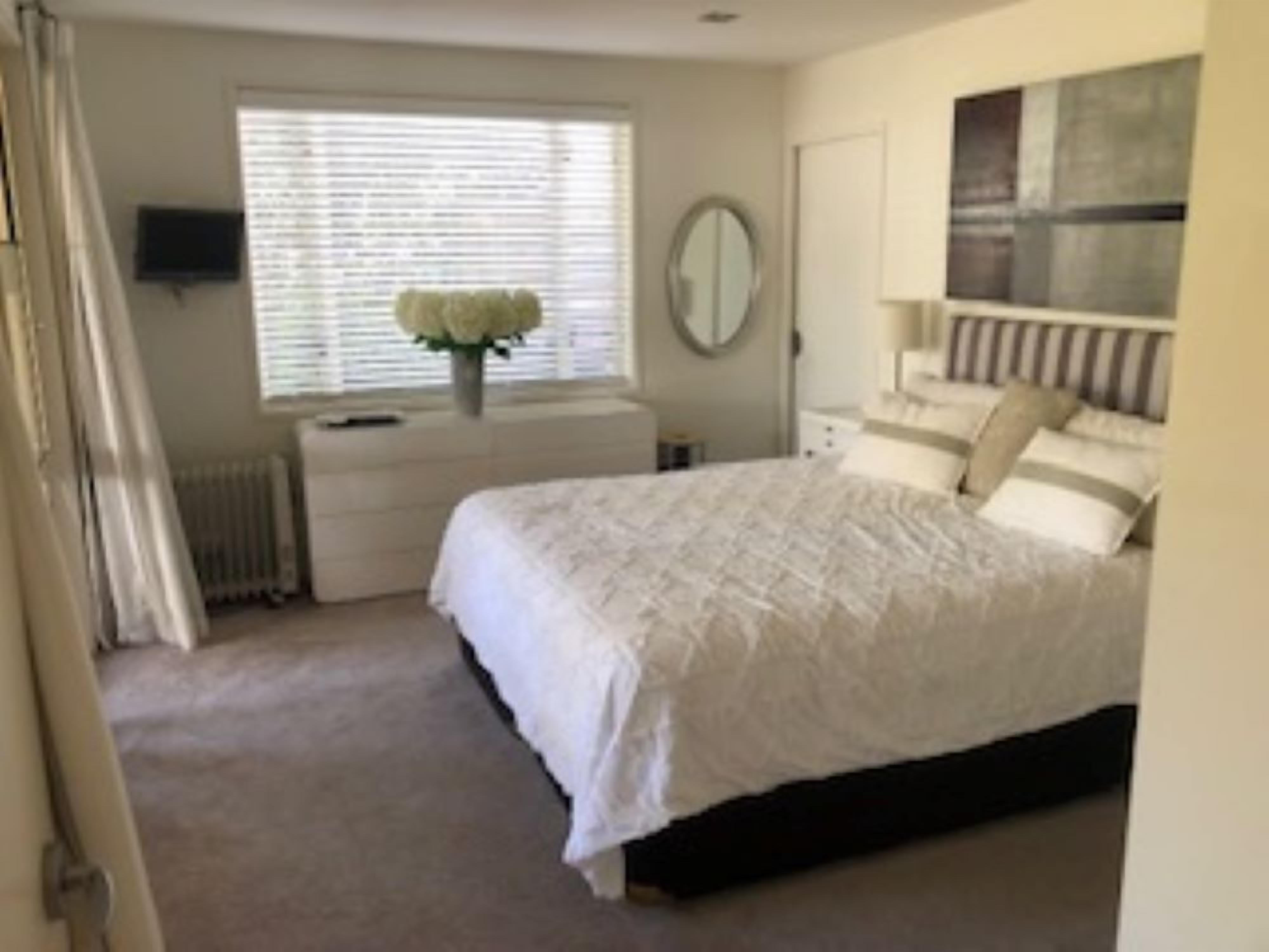Een slaapkamer met een bed, nachtkastje, spiegel, radiator en een raam met jaloezieën.