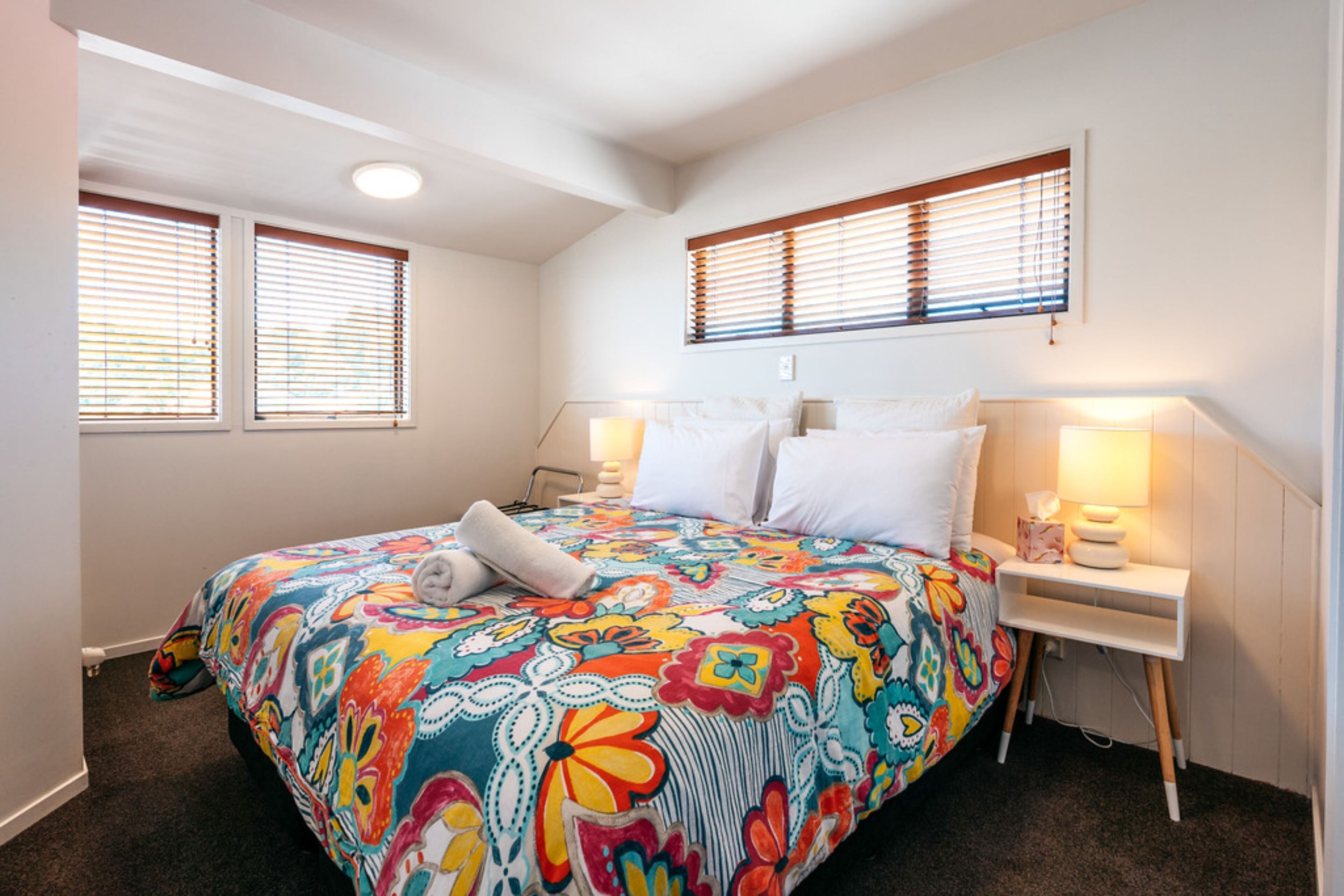 A bedroom with a colorful floral bedspread, two bedside tables with lamps, and a window with blinds.