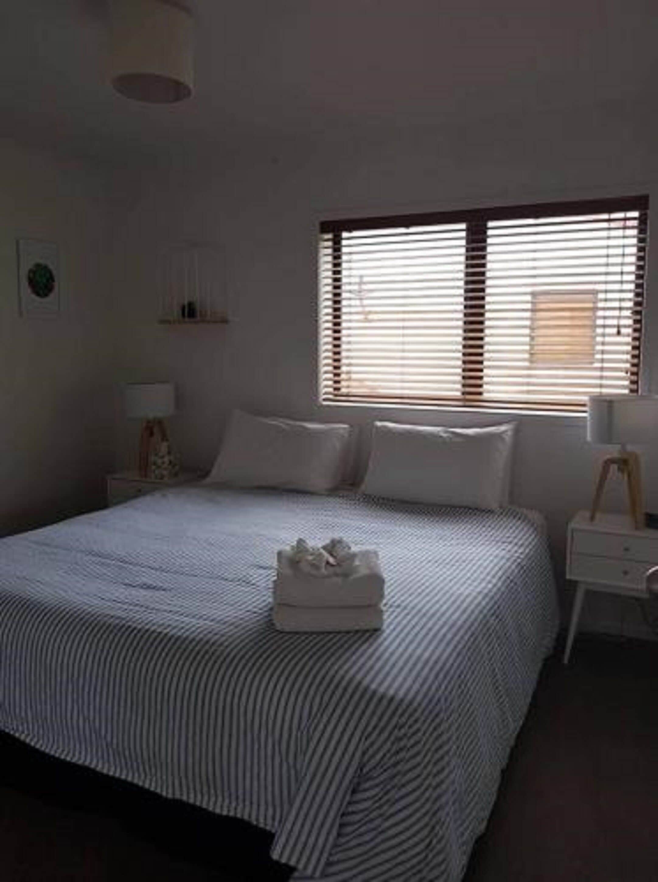 Een slaapkamer met een bed, een nachtkastje met een lamp, een raam met zonwering, en een wandplank.