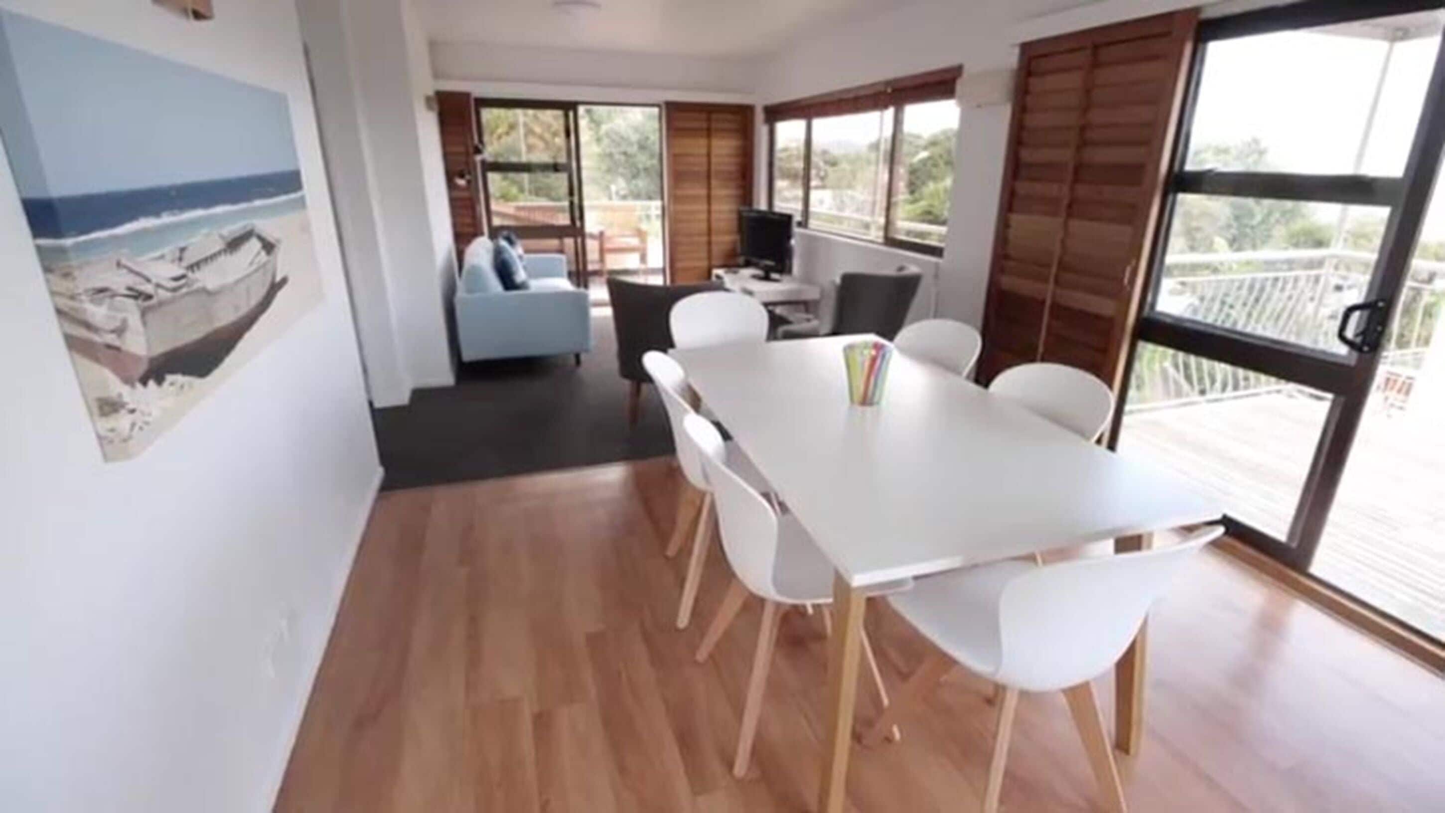 Un comedor con una mesa y sillas blancas, un cuadro en la pared y ventanas grandes con persianas.