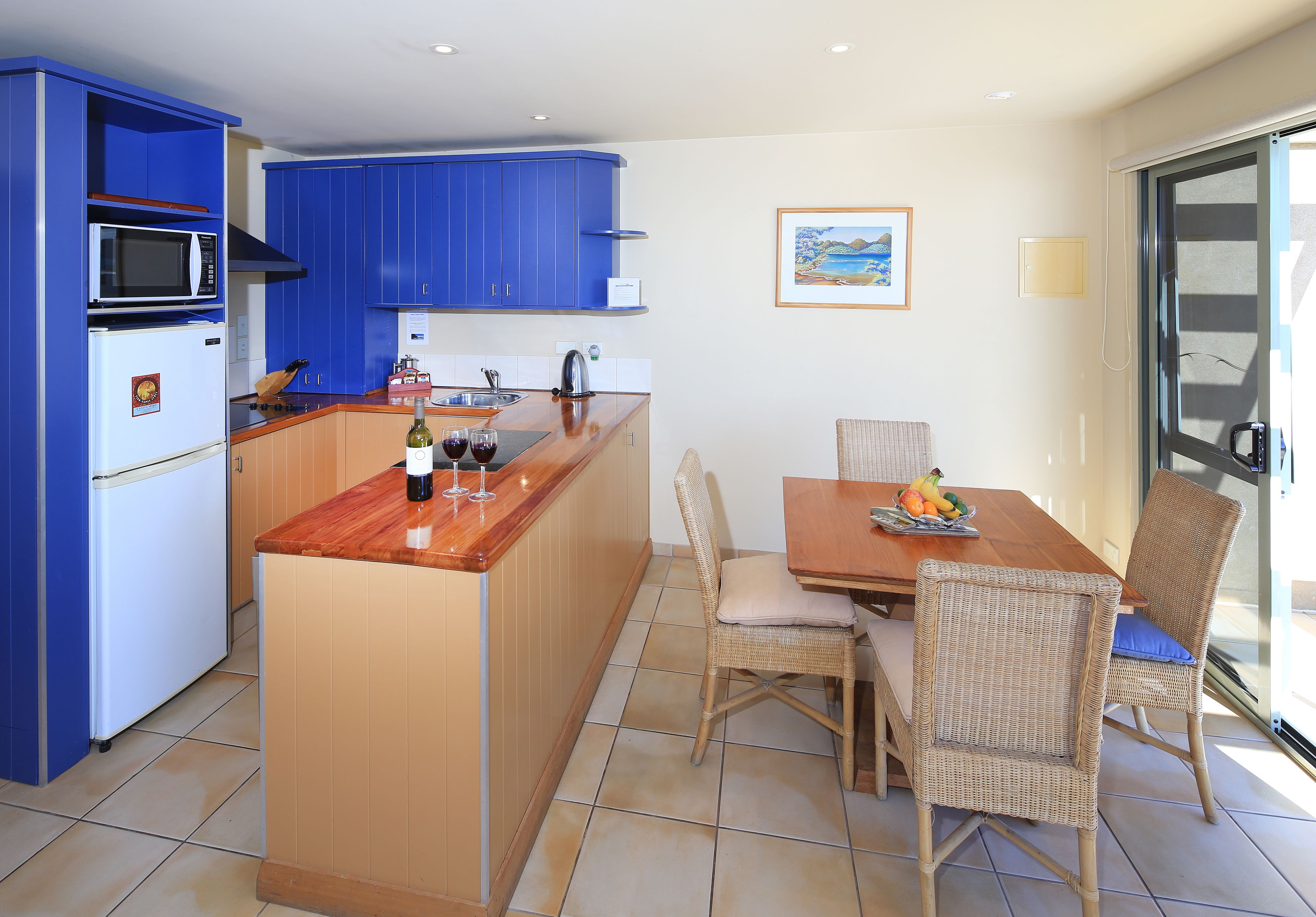 A compact kitchen with blue cabinets, a wooden island, and a dining area with wicker chairs.