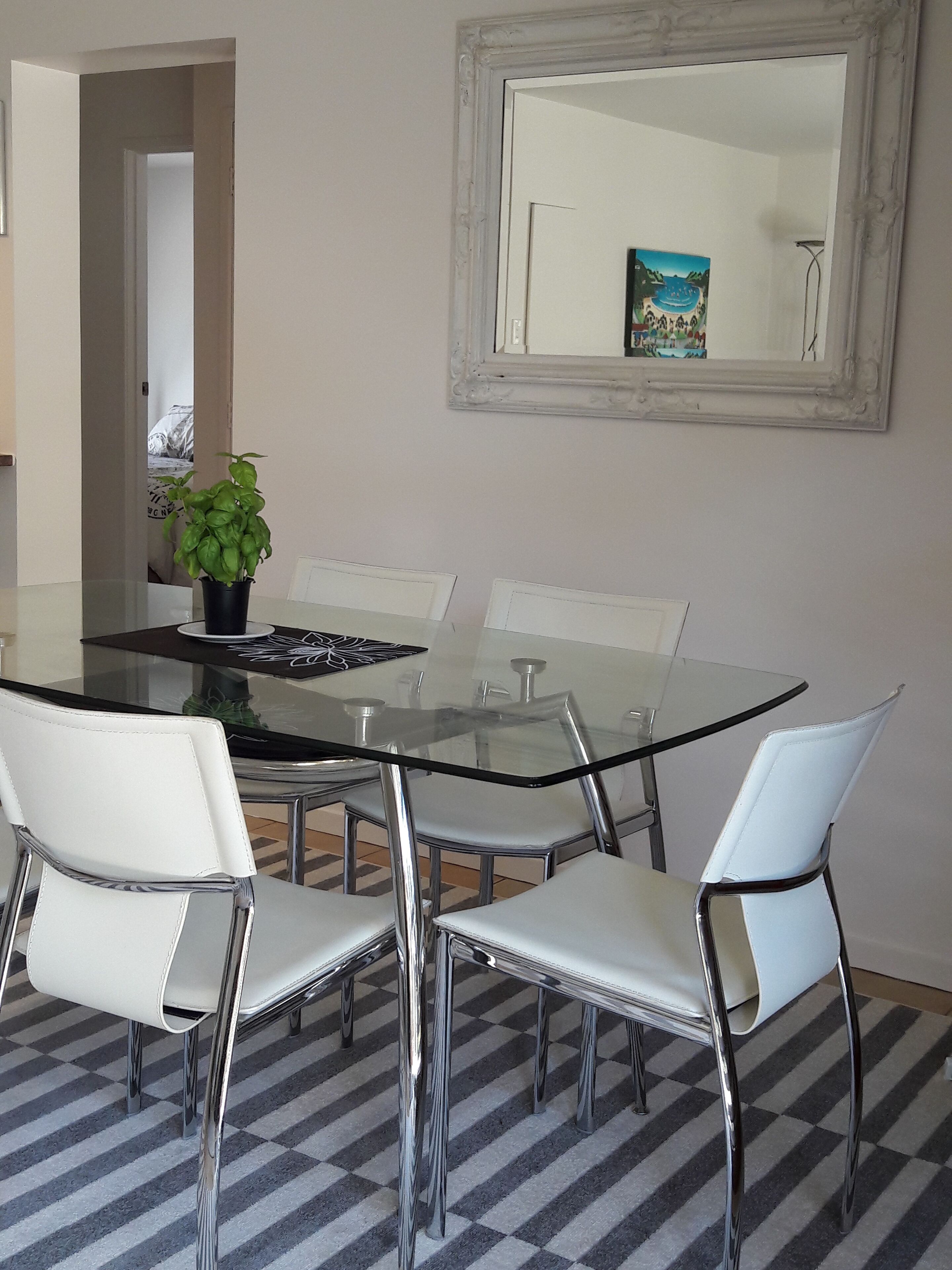 Un comedor con una mesa de vidrio rodeada de sillas blancas, una planta en maceta y un cuadro en la pared.