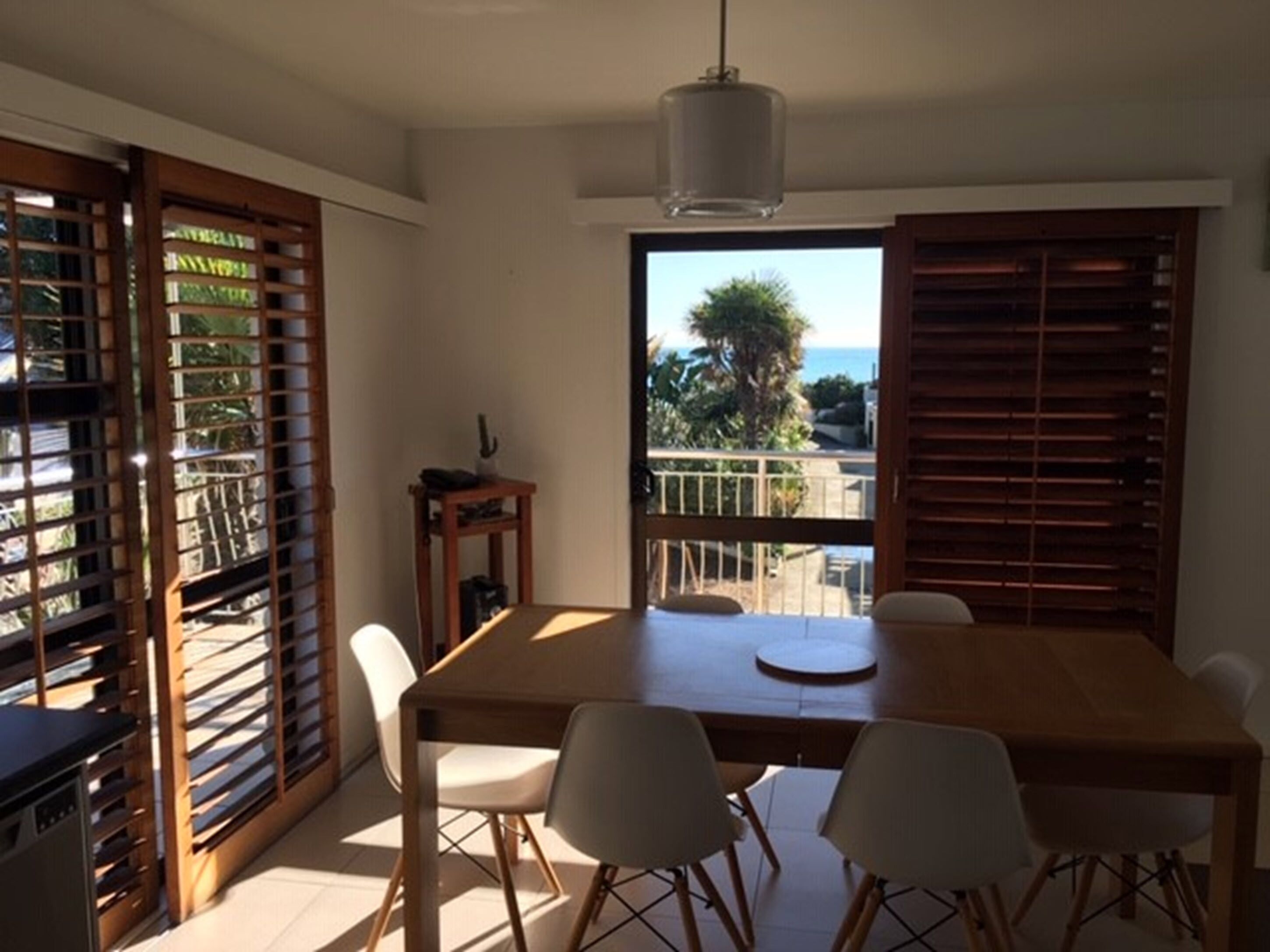 A dining area with a wooden table and white chairs, a small side table, and a view of the outdoors through a large window.