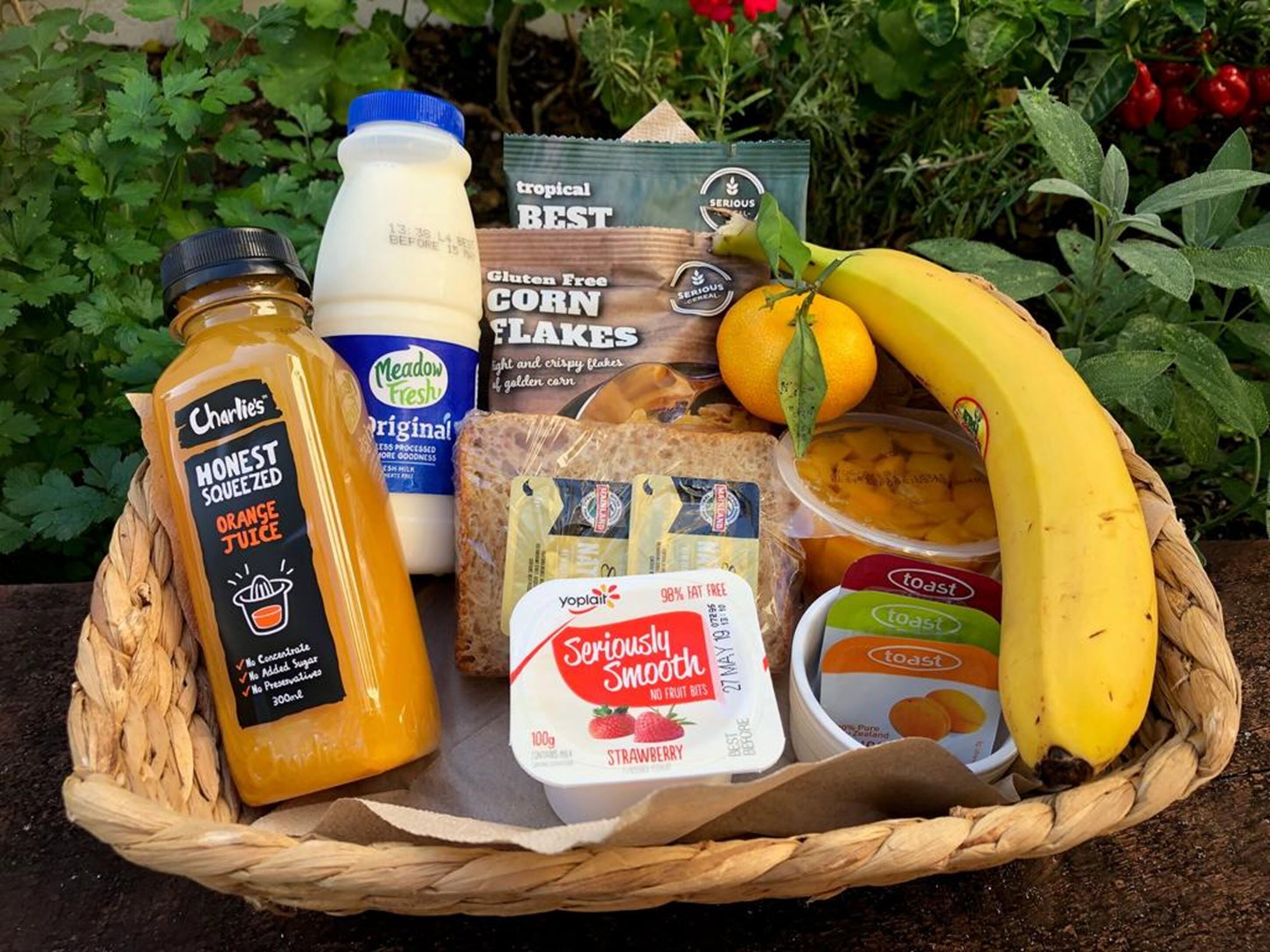 Una cesta con una botella de jugo de naranja, un cartón de leche, una barra de pan, un plátano, un envase de yogur y una caja de copos de maíz.