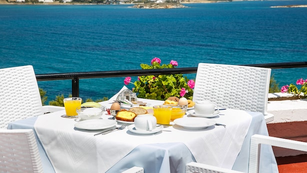 Mesa dispuesta con alimentos para el desayuno, mantel blanco y vista al mar y las colinas.