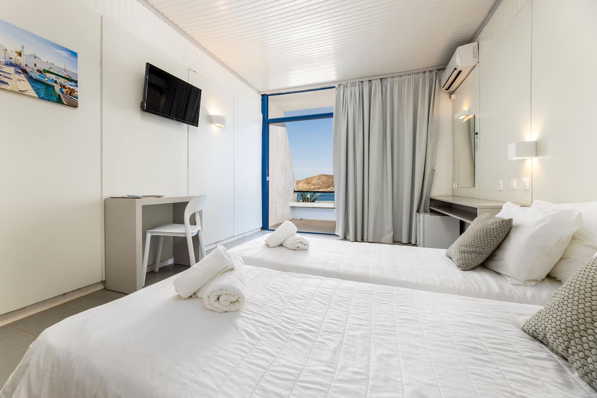 A hotel room with two beds, a desk, a chair, a TV, and a view of the sea.