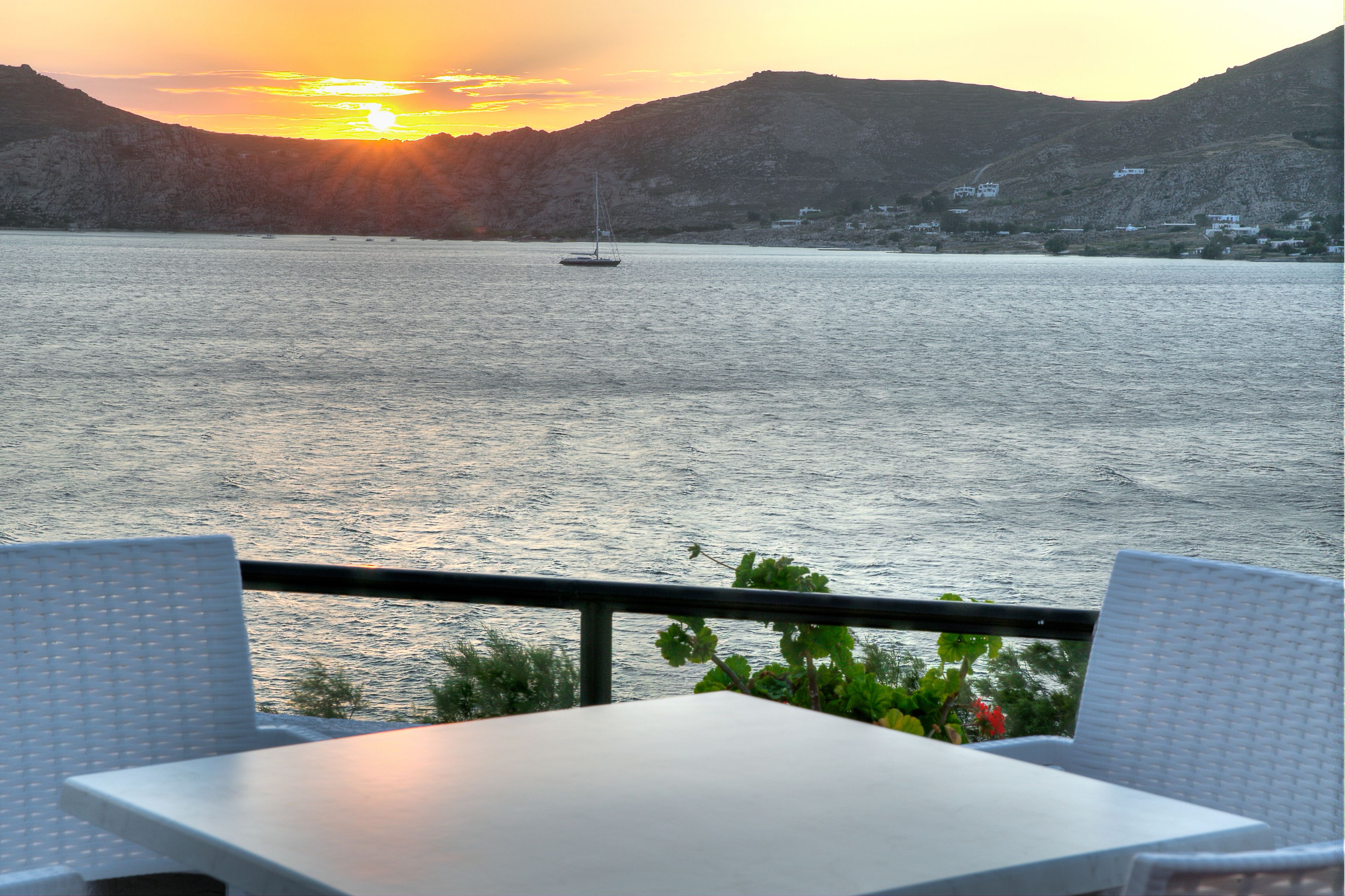 A table and chairs on a balcony overlooking a body of water with a sailboat in the distance and a mountainous landscape in the background.