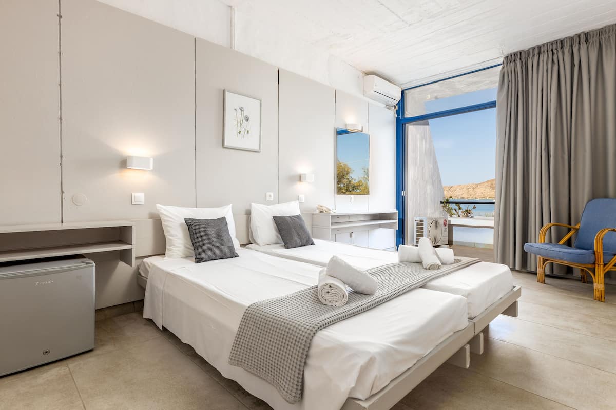 A modern hotel room with two beds, a small kitchenette, a balcony with a view, and a wicker chair.