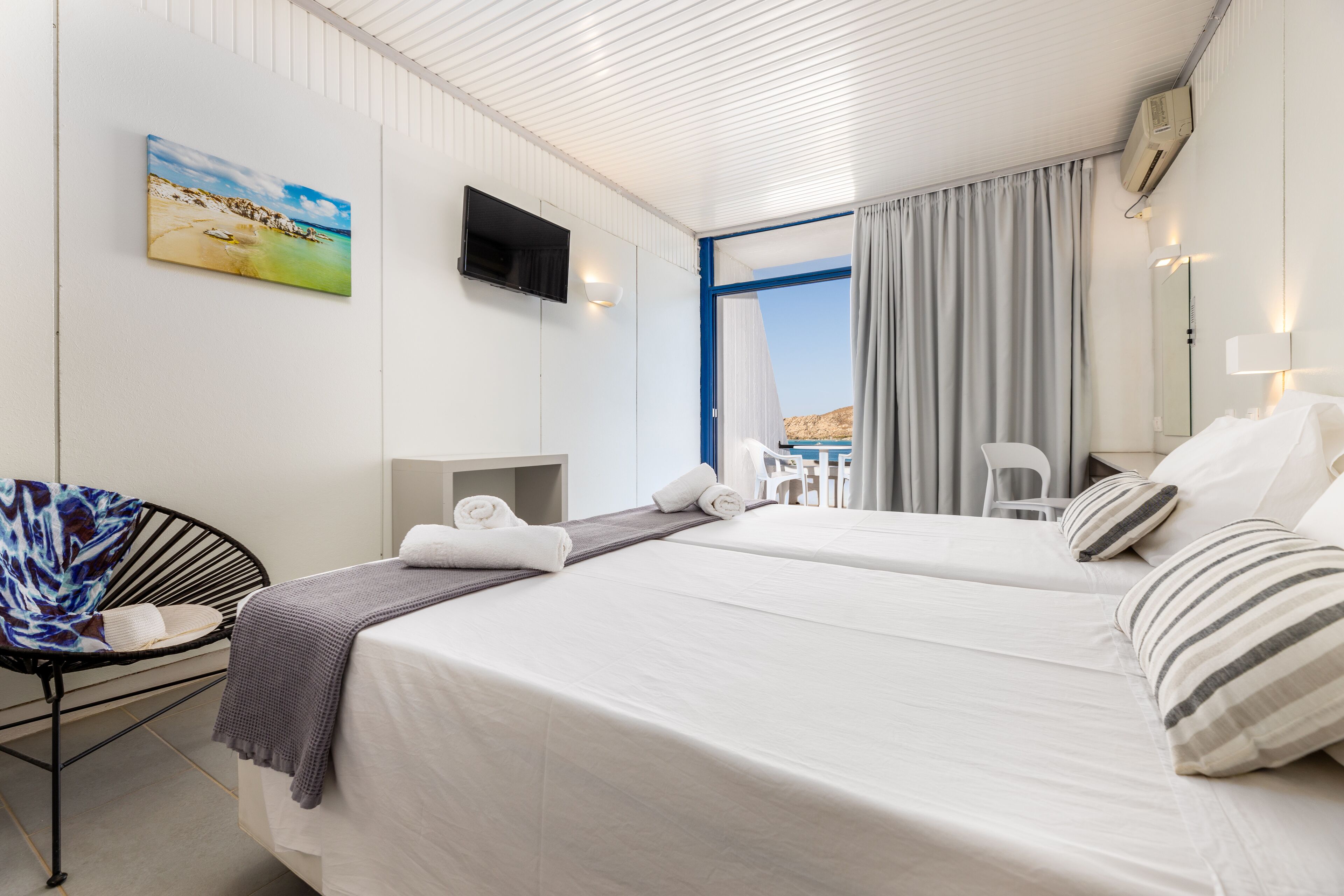 Habitación de hotel con dos camas, un televisor montado en la pared, un cuadro de playa y vistas al exterior a través de un ventanal grande.