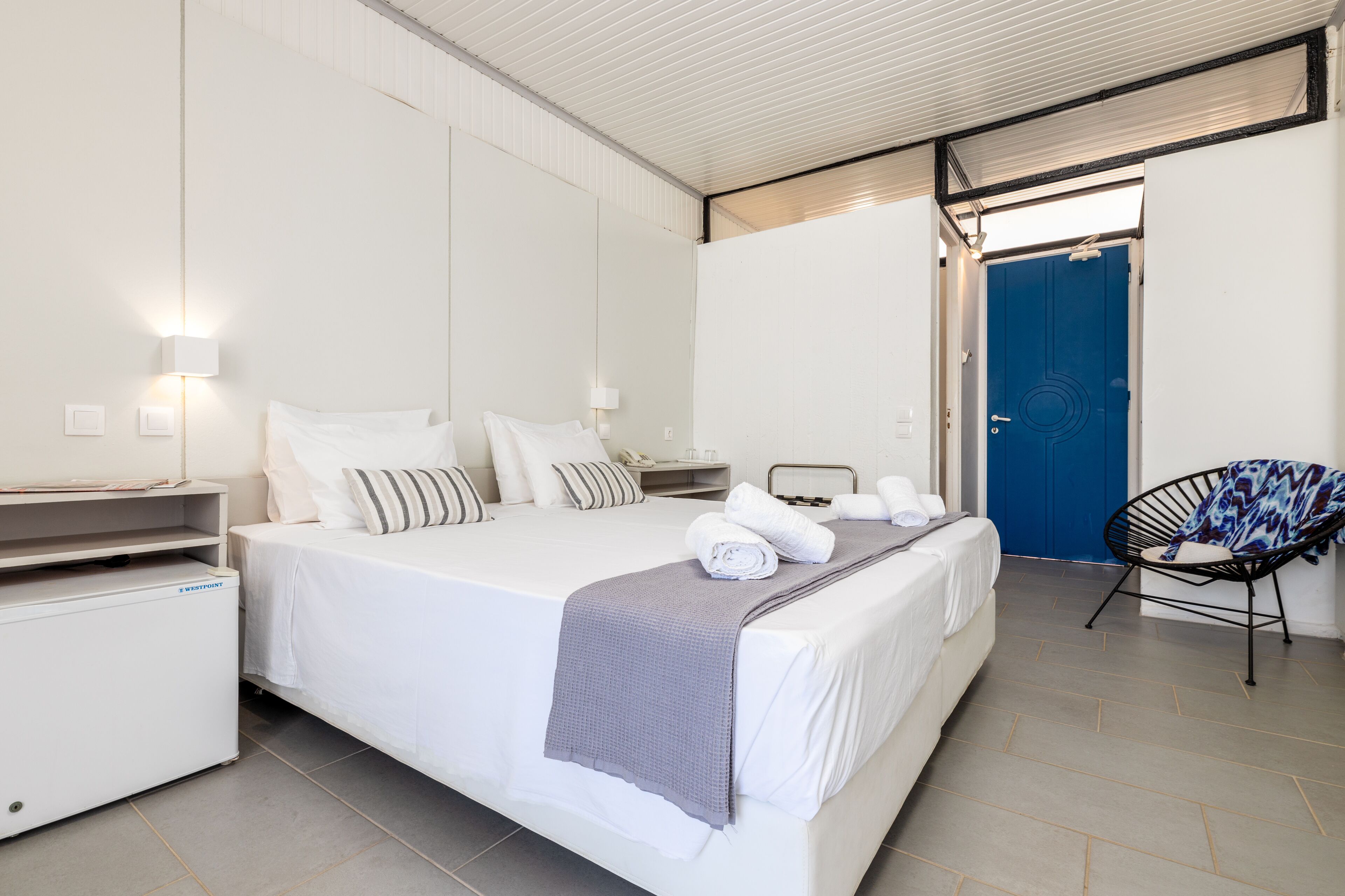 Una habitación de hotel con cama, mesitas de noche, una silla y la puerta del baño.