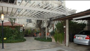 A covered outdoor area with a car parked under a pergola, surrounded by greenery and a building in the background. - Grand Hotel Balbi (Mendoza)