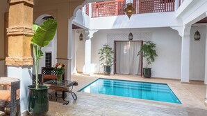 A pool area with a table and chairs, potted plants, and a balcony with red railings.