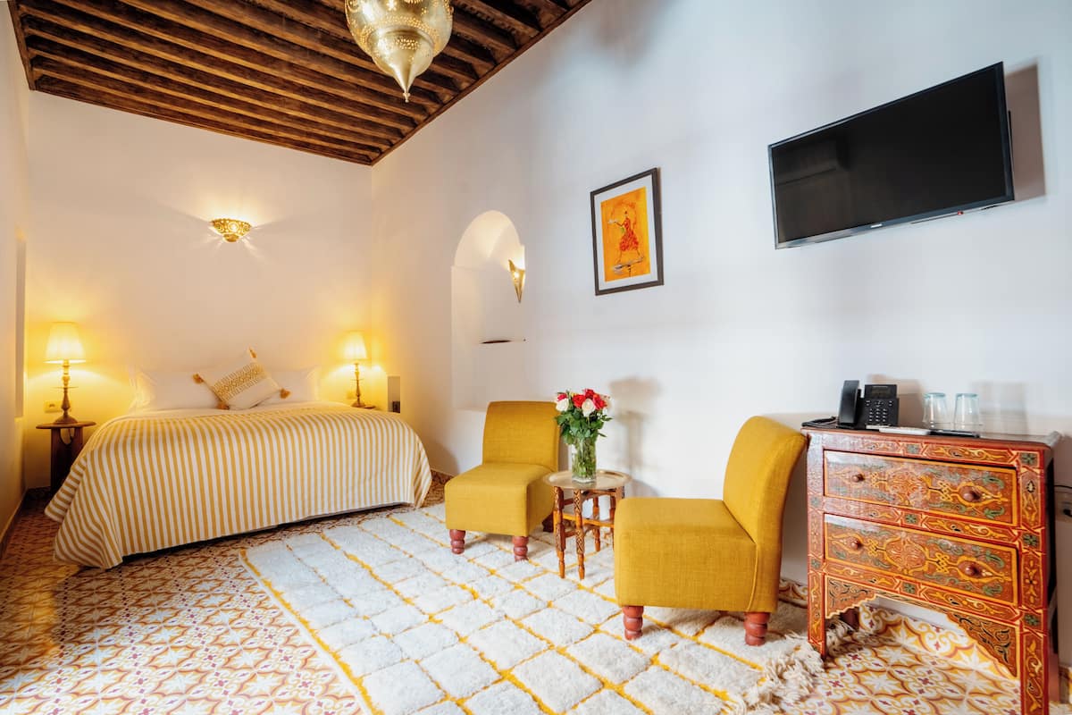 A hotel room with a bed, two yellow armchairs, a small table with a vase, a wooden dresser, a flat-screen TV, and a patterned rug.