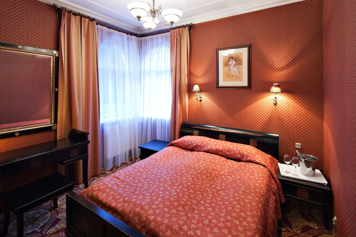 A hotel room with a bed, a desk, a chair, a nightstand with a lamp, a picture on the wall, and a window with curtains.