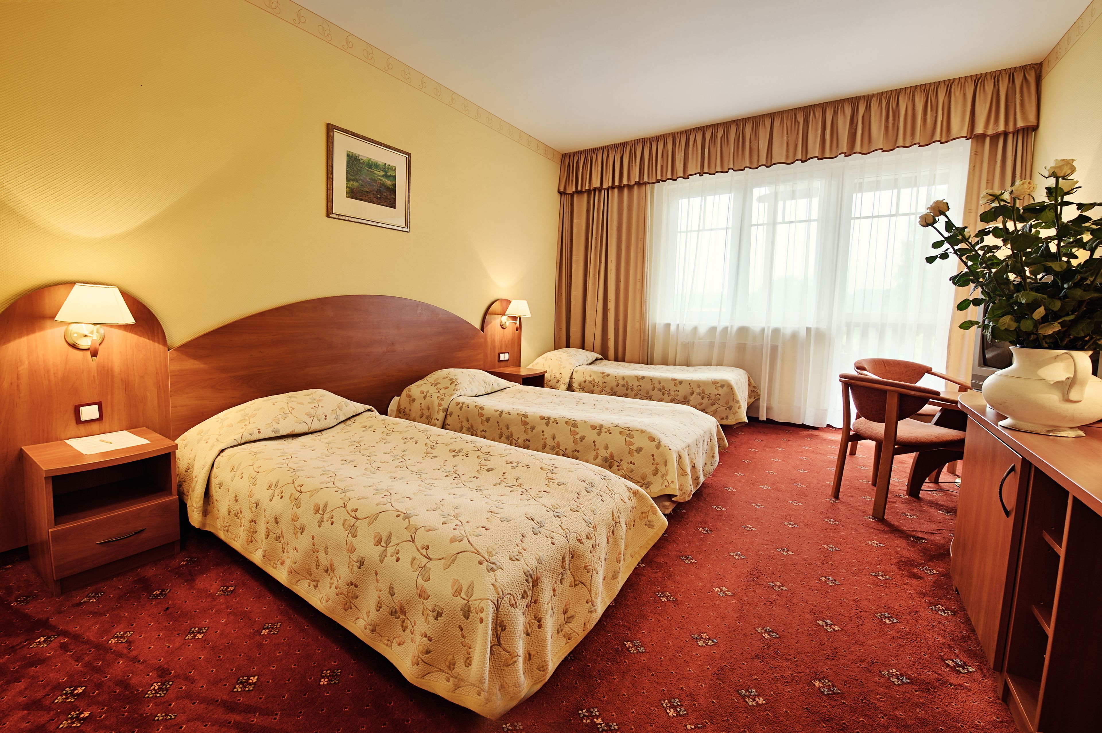 A hotel room with two beds, a desk, a chair, a lamp, a picture on the wall, and a window with curtains.