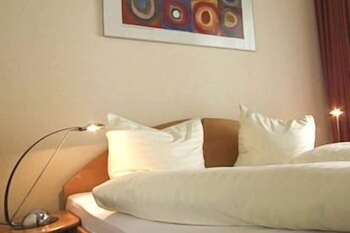 Habitación de hotel con cama, almohadas, mesita de noche, lámpara y un cuadro en la pared.