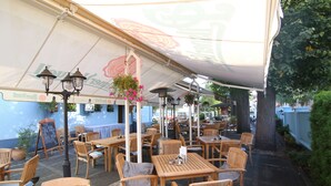 An outdoor cafe with wooden tables and chairs, hanging flower baskets, and a large umbrella providing shade. - Agh Hotel (Roznov Pod Radhostem)