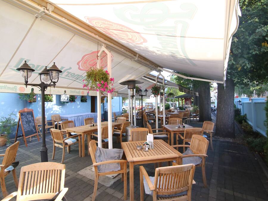 An outdoor cafe with wooden tables and chairs, hanging flower baskets, and a large umbrella providing shade.