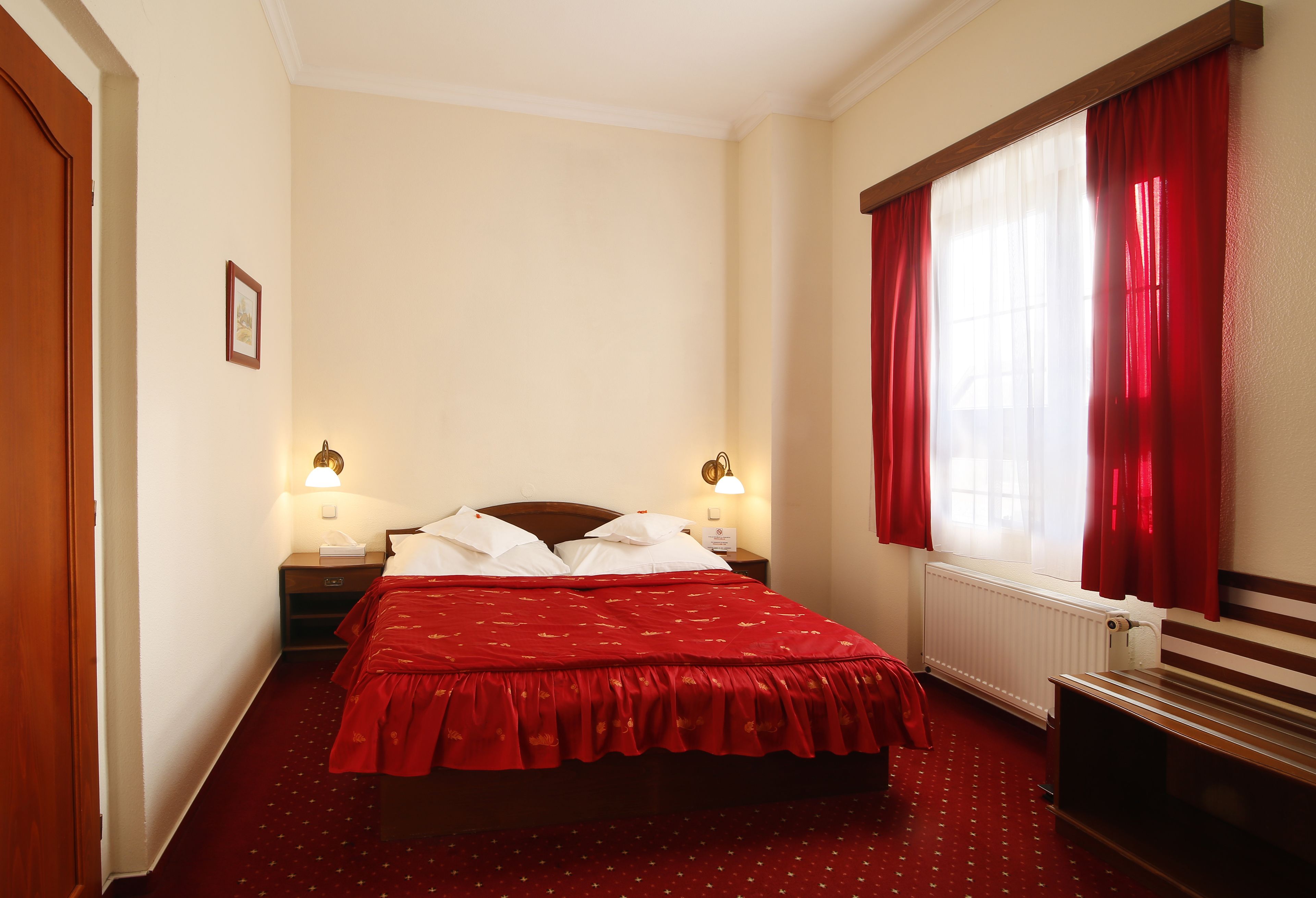 Habitación de hotel con cama, dos mesitas de noche, un banco, una ventana con cortinas rojas y un radiador.