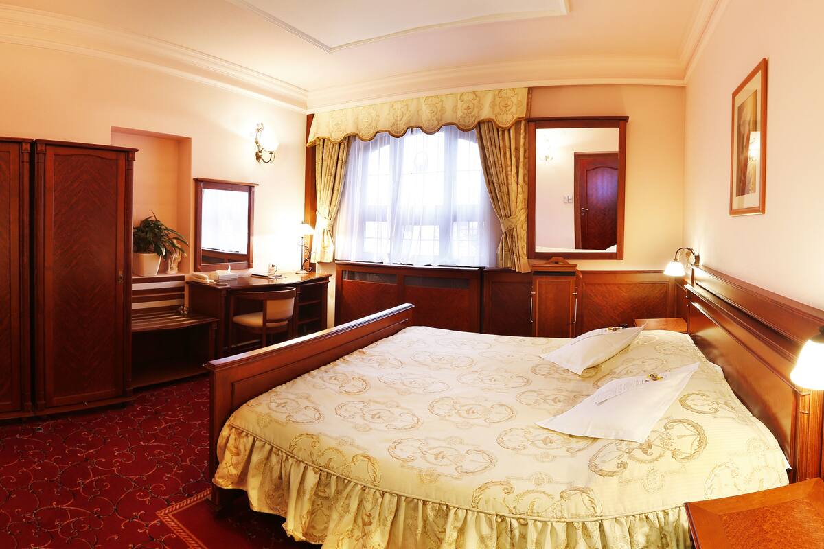 A hotel room with a large bed, a desk, a chair, a wardrobe, and a window with curtains.