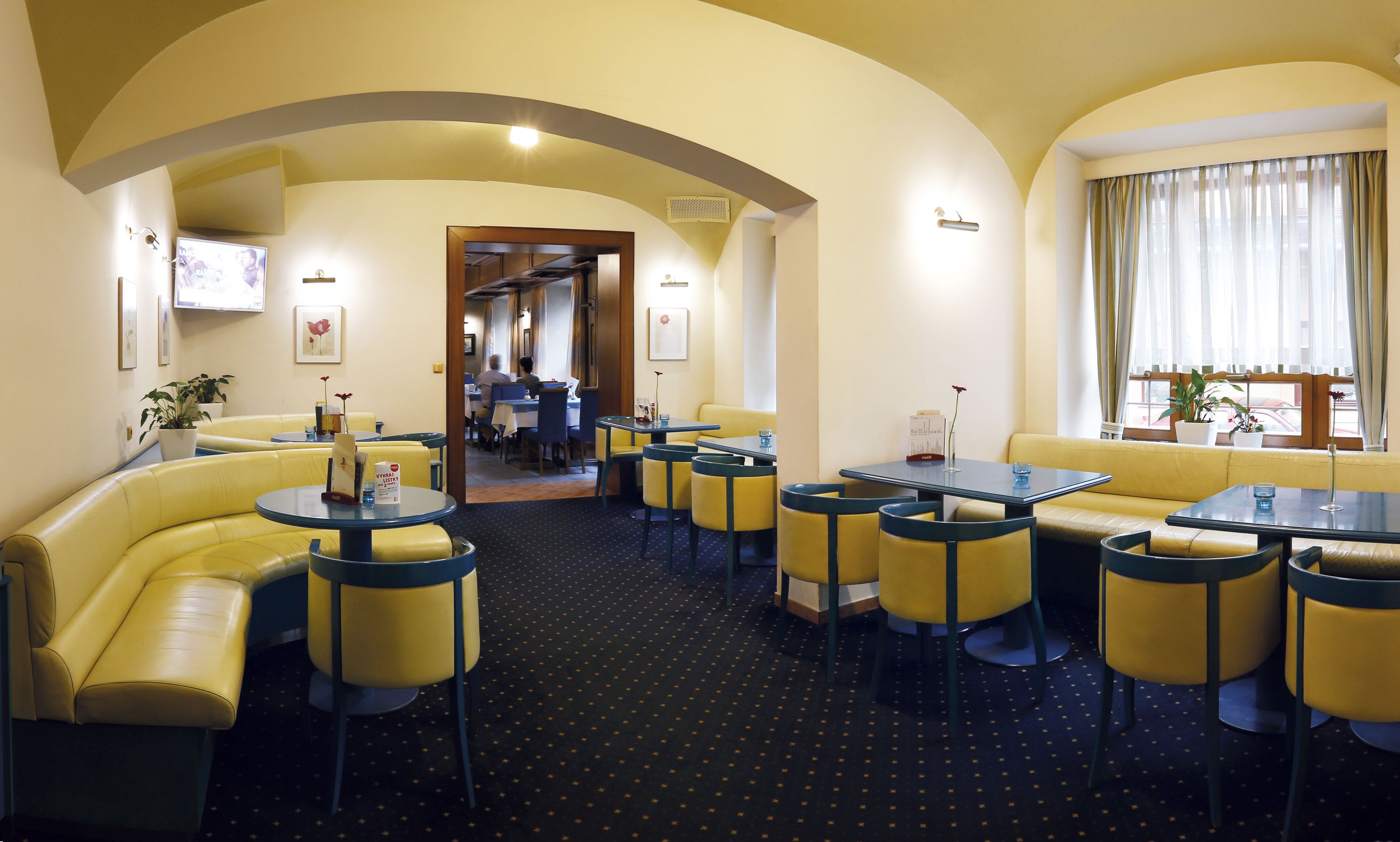 Un restaurante con asientos tipo banqueta amarillos y mesas redondas, con alfombra a puntos negros y techo abovedado.