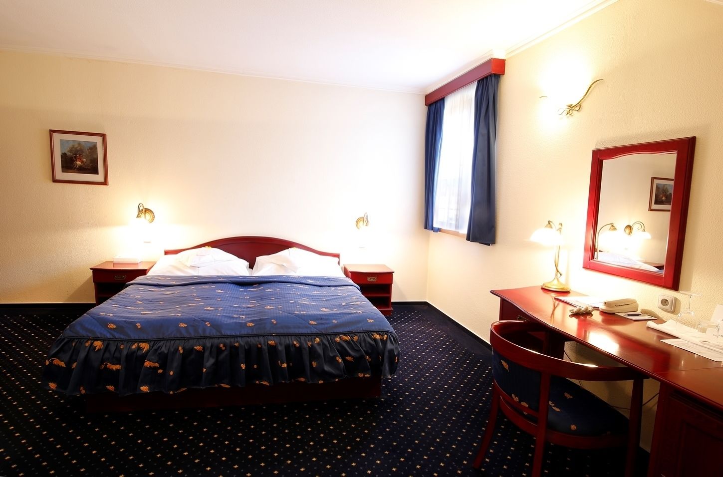 Une chambre d’hôtel comprenant un lit, des tables de chevet, un bureau, une chaise, un miroir et une fenêtre avec des rideaux.