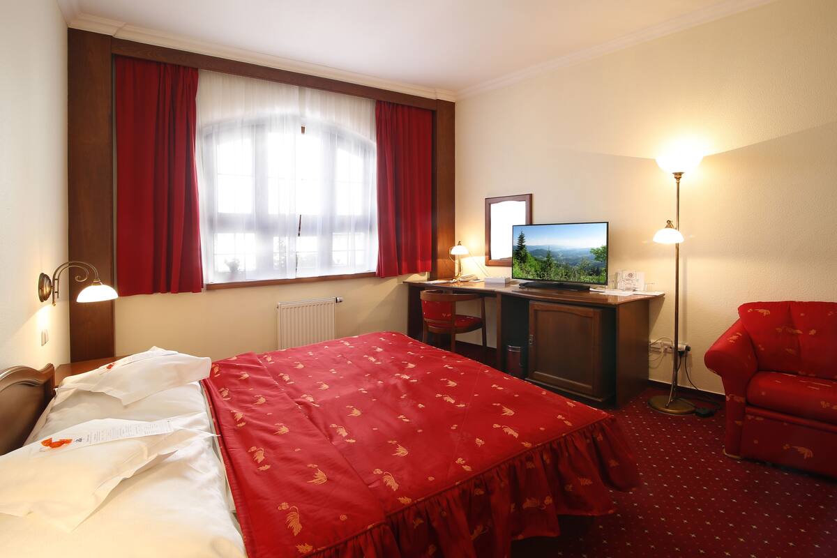 A hotel room with a bed, a desk with a television, a red chair, and a floor lamp.