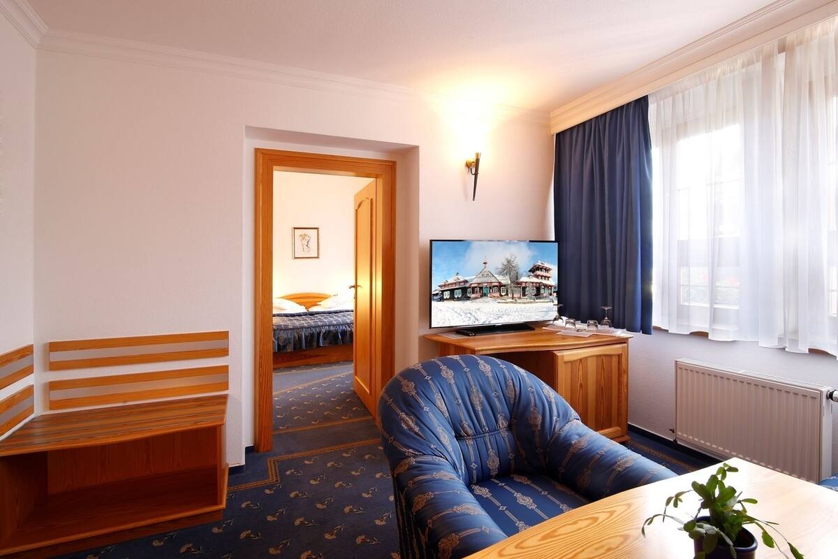 A hotel room with a bed, a TV, a blue armchair, a wooden side table, and a window with curtains.