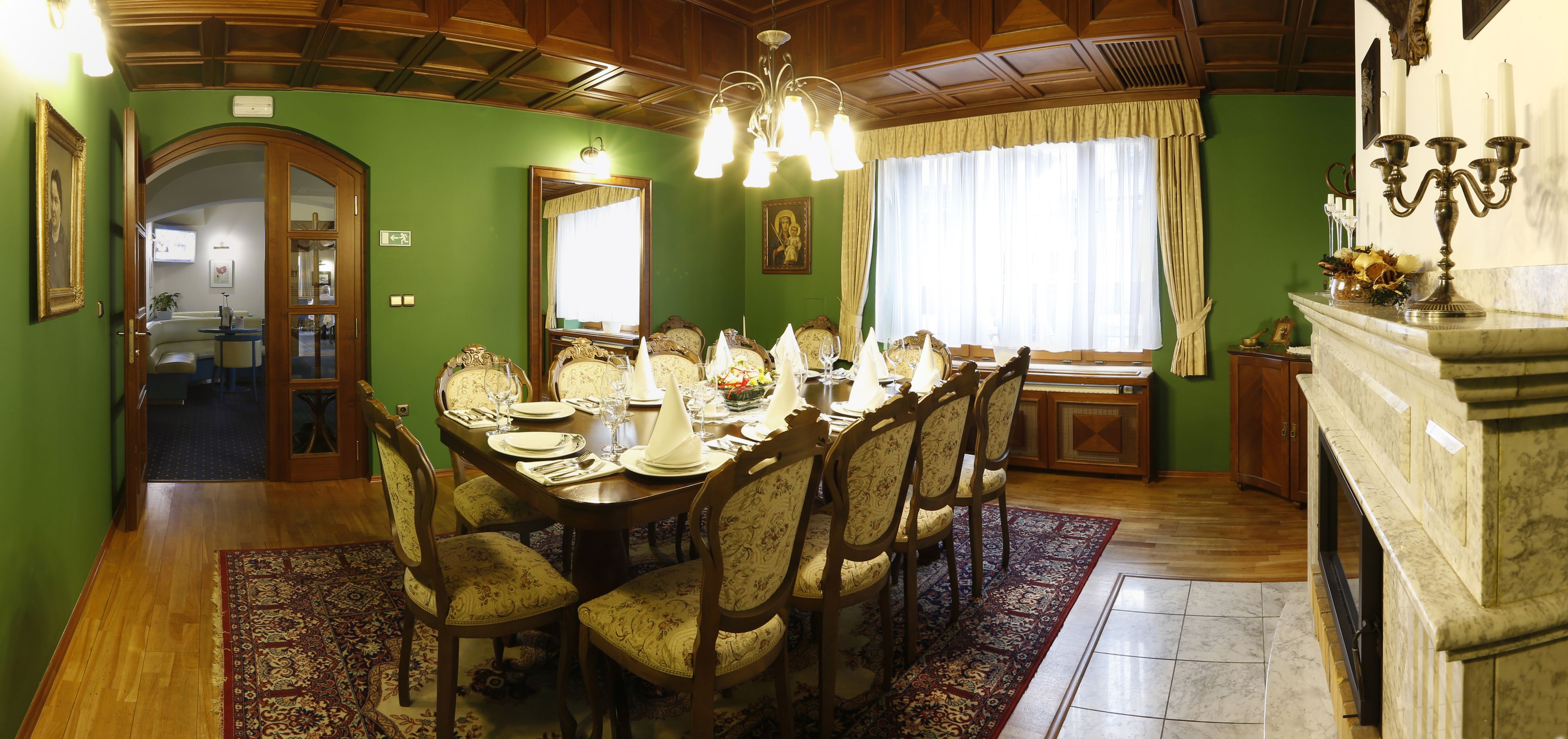 A dining room with a wooden table set for six, surrounded by upholstered chairs. A chandelier hangs from the ceiling, and a fireplace is visible on the right.