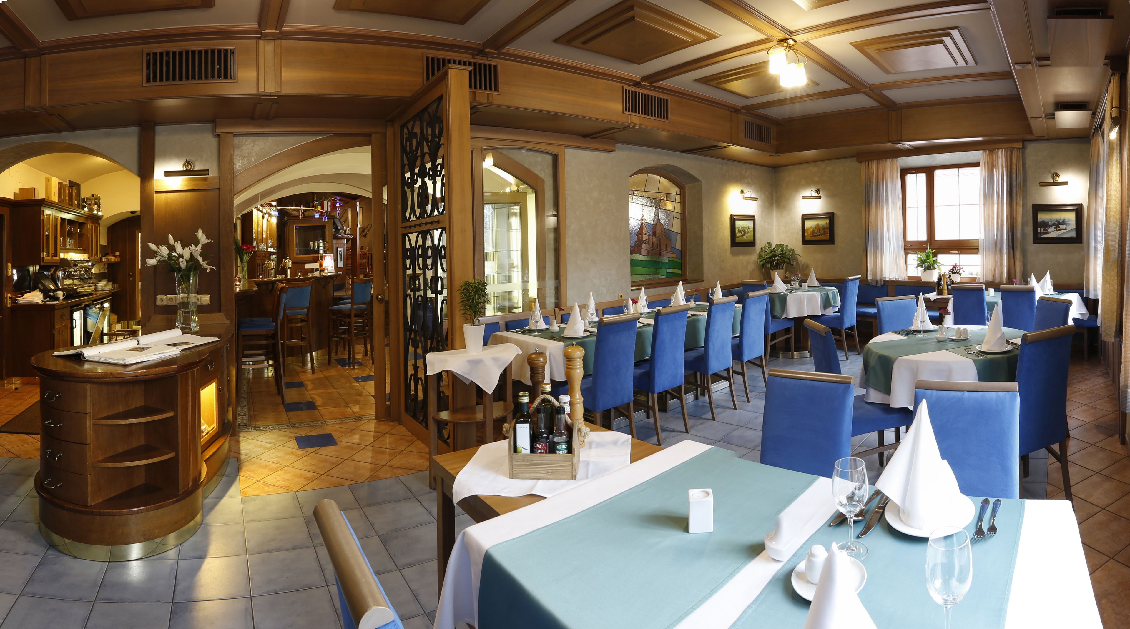 A restaurant with a classic European interior, featuring wooden beams, arched doorways, and blue upholstered chairs. Tables are set with white tablecloths and silverware.