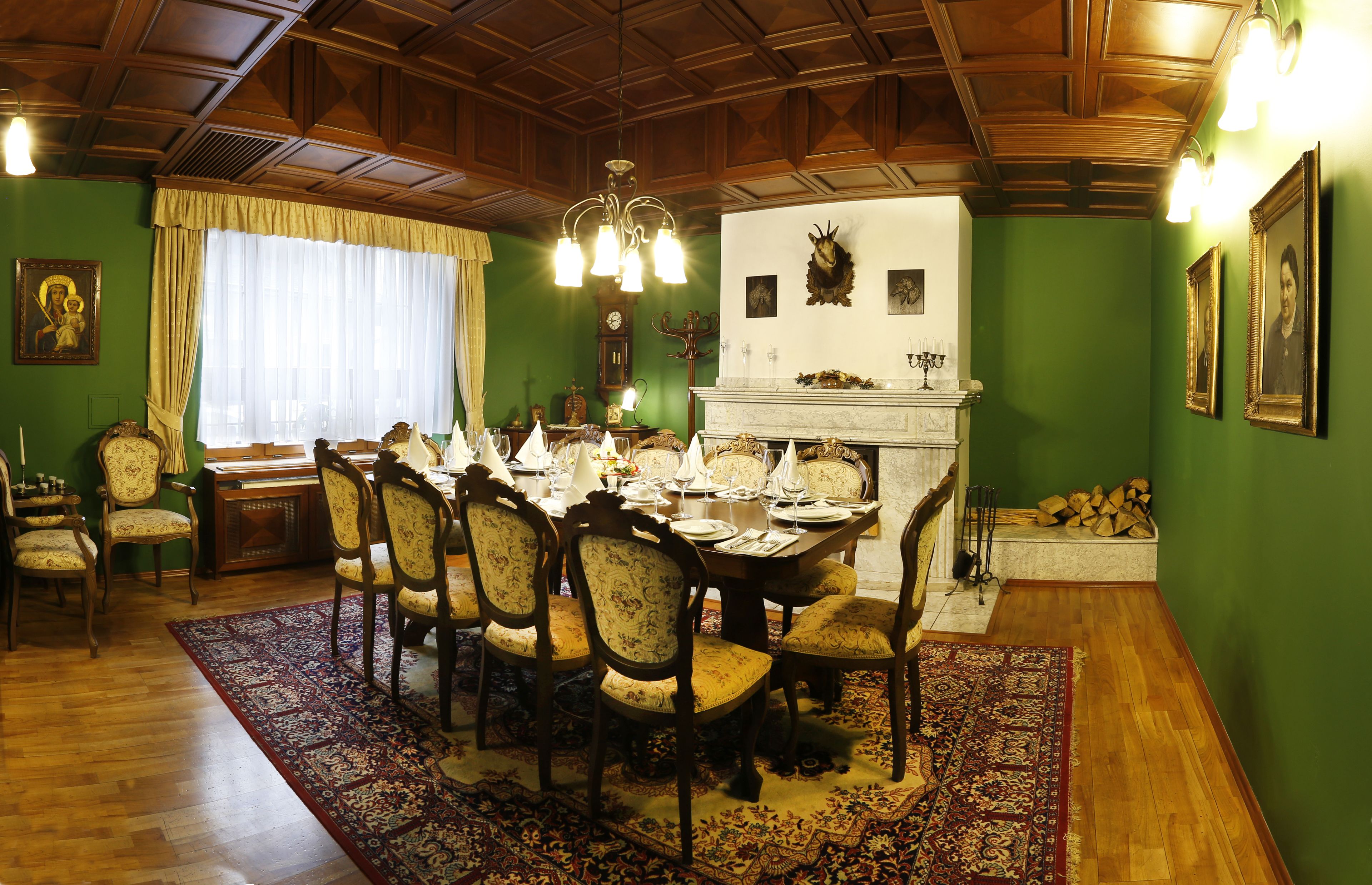 A dining room with a wooden table set for six, surrounded by chairs. A chandelier hangs from the ceiling, and a fireplace is visible in the background.
