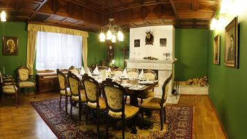A dining room with a wooden table set for six, surrounded by chairs. A chandelier hangs from the ceiling, and a fireplace is visible in the background.