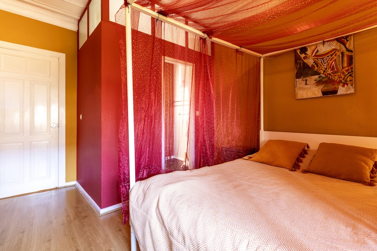 A bedroom with a bed, a window with curtains, a door, and a painting on the wall.