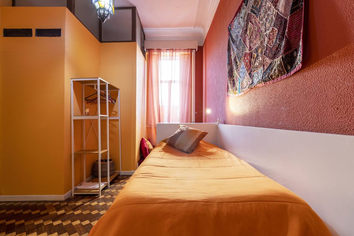 A room with a bed, a wardrobe, a window with curtains, and a wall with a colorful tapestry.