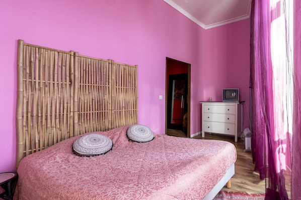 A bedroom with a pink wall, a bamboo headboard, a bed with patterned bedding, a small white dresser, and a television on a nightstand. - Porto Riad Guest House (Porto)