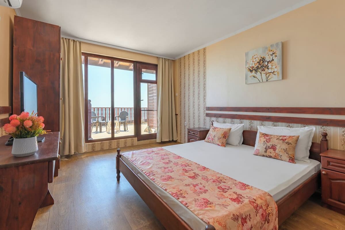 A hotel room with a large bed, a desk with a vase of flowers, a painting on the wall, and a balcony with chairs.