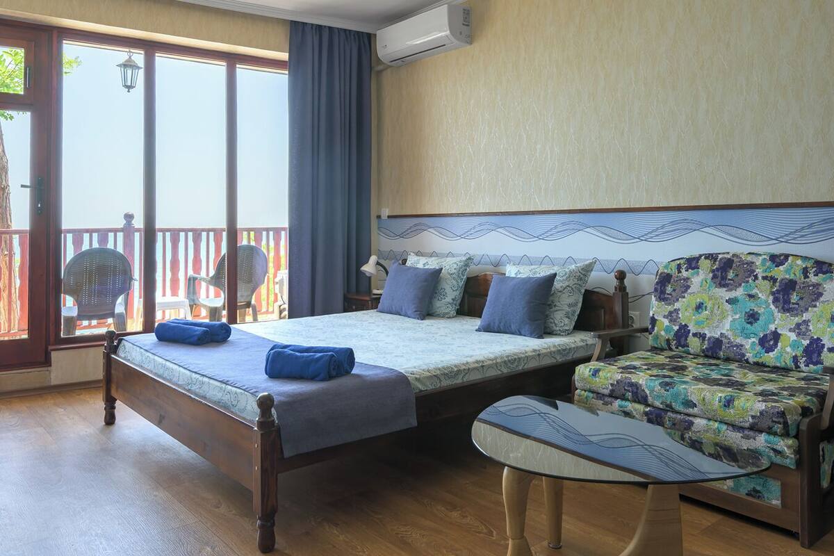 A bedroom with a bed, a sofa, and a chair. There is a balcony with chairs and a table.