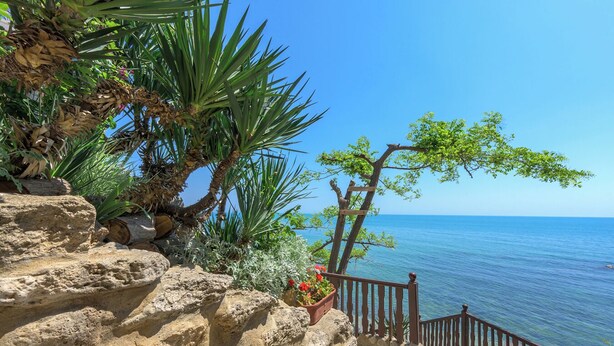 Vista costera con una escalera de madera que lleva a un balcón con vistas al mar, rodeado de rocas y vegetación.