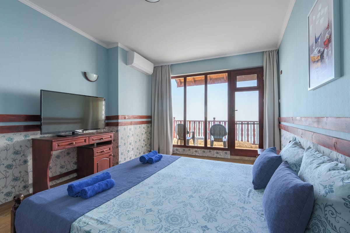 A hotel room with a bed, a desk with a flat-screen TV, a balcony with chairs, and a view of the outside.